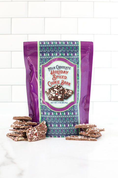 Milk Chocolate Holiday Spiced Cookie Bark with festive packaging and sprinkled chocolate pieces on a white countertop.