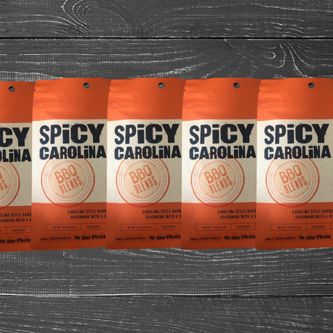 Five packs of Spicy Carolina BBQ Seasoning on a wooden surface, showcasing vinegar and pepper-based BBQ blends for grilling and marinades.