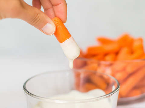 Person dipping carrot stick in creamy white sauce with bowl of carrots in background.